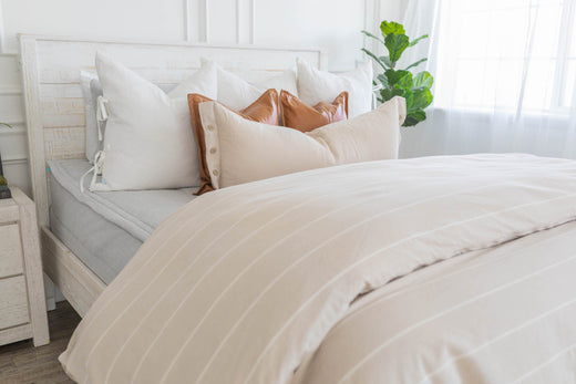 A bed made with light zipper bedding with a duvet folded at the foot of the bed and an assortment of throw pillows.