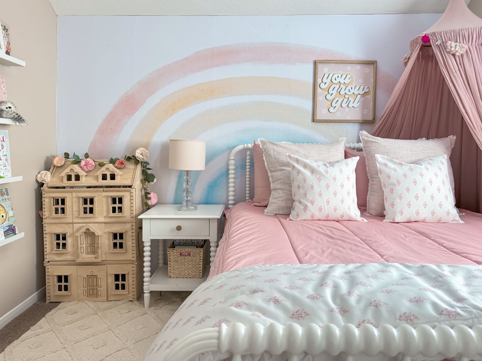 
      A cozy, pastel-themed girl's bedroom featuring pink Bed-dys bedding, patterned pillows, a nightstand, a wooden dollhouse adorned with pink flowers, and a whimsical rainbow mural on the wall.
    
