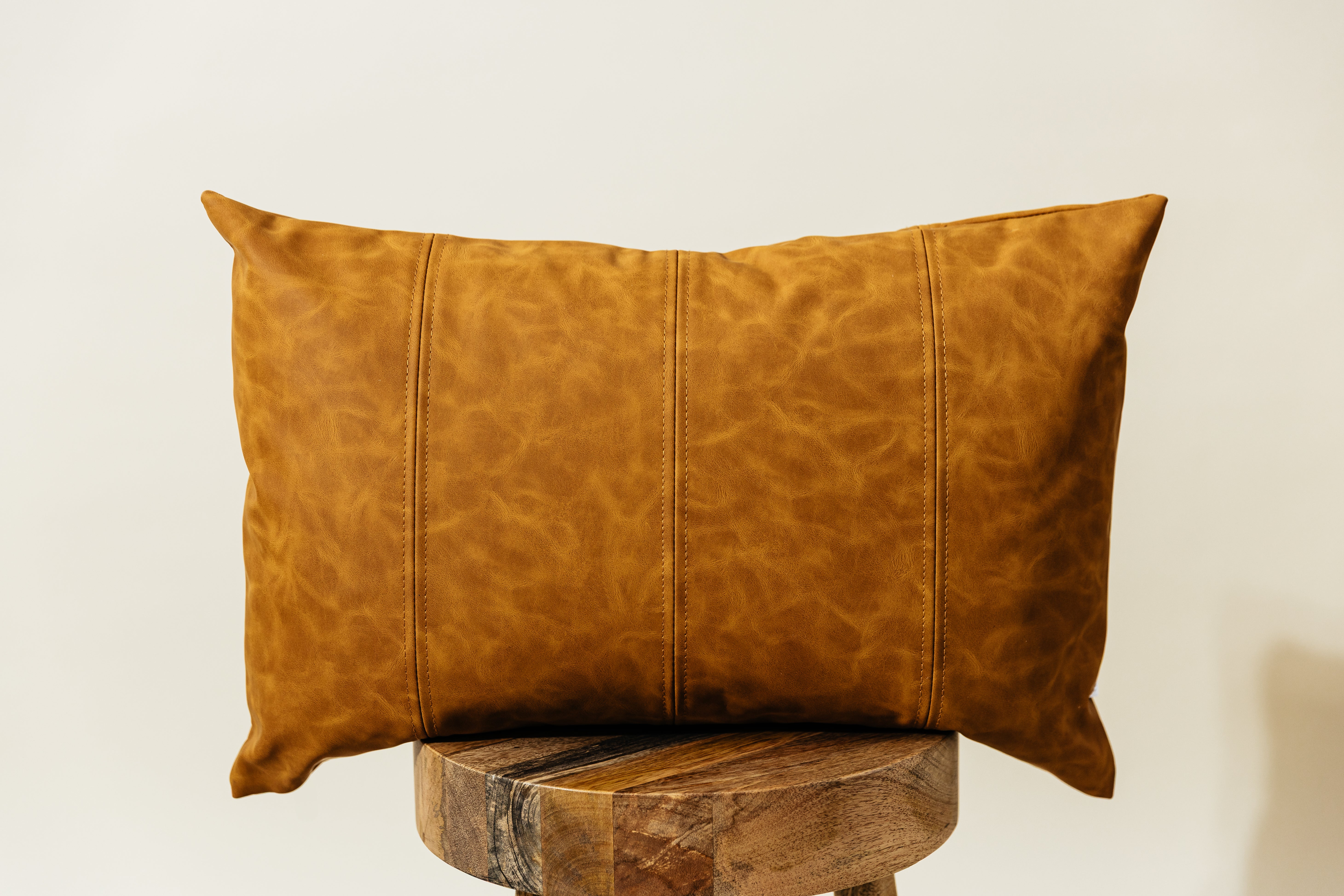 A faux leather lumbar pillow sitting on a wood stool in front of a white backdrop.