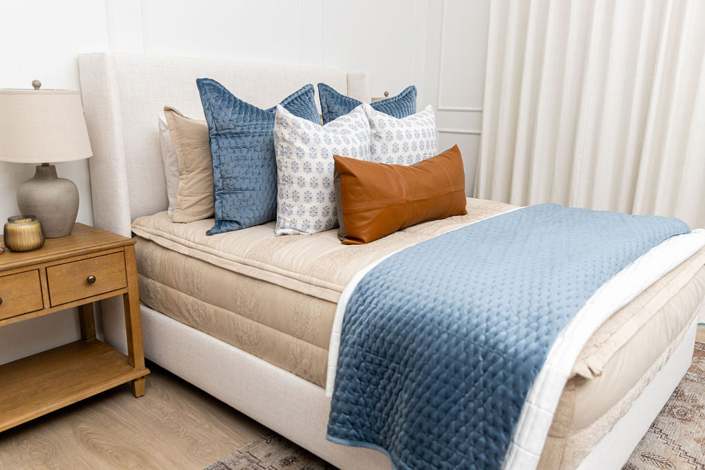 A queen bed made with tan zipper bedding and an assortment of blue, leather, and white bedding accessories.