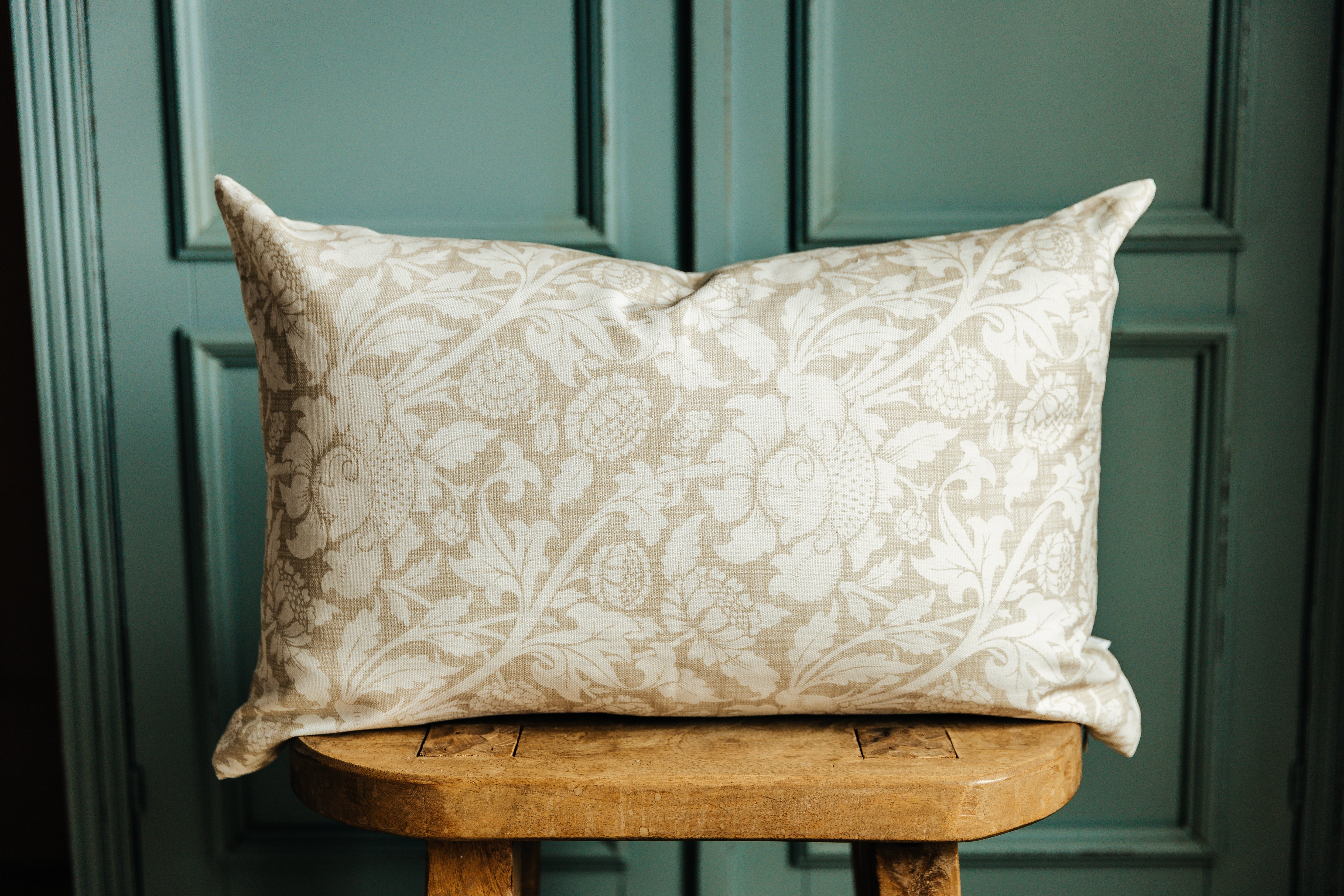A floral lumbar pillow on a wood stool in front of a green wall.